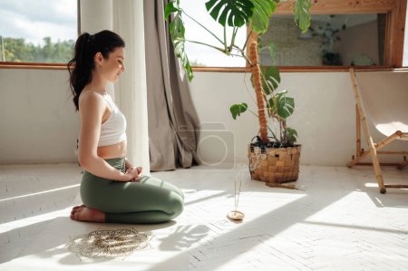 Téléchargez les photos : Femme calme et spirituelle assise à genoux sur un tapis de yoga blanc avec les yeux fermés dans une pièce lumineuse. Femme attrayante habillée de vêtements de sport confortables méditant sur le sol. - en image libre de droit