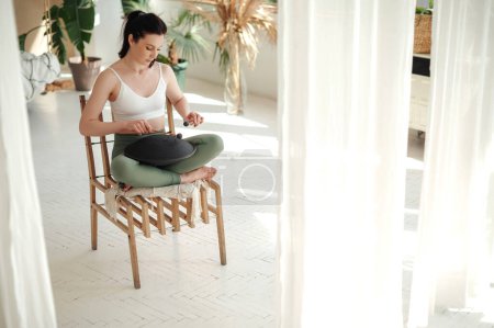 Foto de Ajuste mujer morena sentada en silla en pose de loto y tocando en tambor de lengua de acero. Adorable hembra relajante para calmar y espiritual. Concepto de paz e inspiración. - Imagen libre de derechos