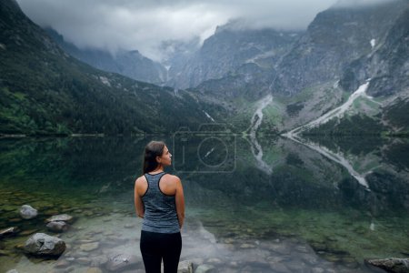 Téléchargez les photos : Vue arrière d'une jeune femme en tenue de sport regardant les collines verdoyantes et les montagnes dans des nuages sombres sur le lac Marine Eye, High Tatras, Zakopane, Pologne. Foggy day - en image libre de droit