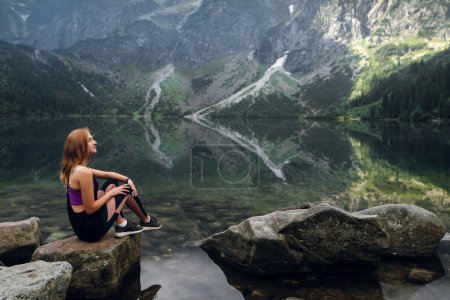Téléchargez les photos : Jeune femme blonde en forme et sportive en tenue de sport assise sur le rocher sur le rivage et admirant une belle vue sur les collines verdoyantes et les montagnes sur le lac Morskie Oko, Hautes Tatras, Zakopane, Pologne. - en image libre de droit