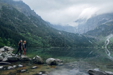 Téléchargez les photos : Deux femmes, amies debout sur la rive pierreuse du lac Moskie Oko avec vue panoramique sur les montagnes avec nuages et brouillard. Rysy mountains, Tatras. Pologne, Slovaquie. Vue arrière. Concept de voyage et de randonnée - en image libre de droit