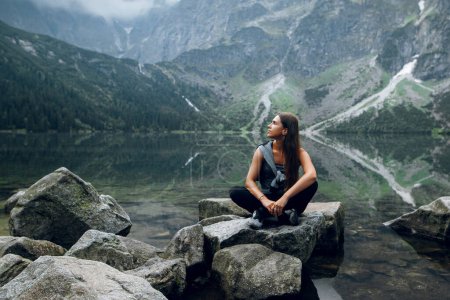 Téléchargez les photos : Belle jeune femme brune en tenue élégante assise sur les rochers, pierres sur le rivage et admirant une belle vue sur les collines verdoyantes et les montagnes sur le lac Morskie Oko, Hautes Tatras, Zakopane, Pologne - en image libre de droit