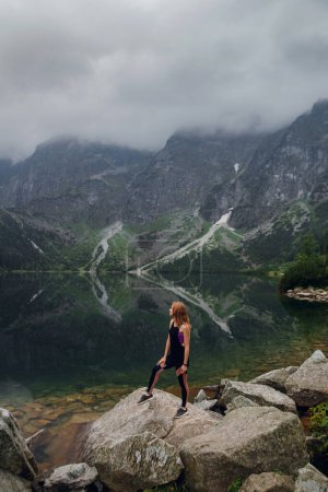 Téléchargez les photos : Jeune belle femme blonde en tenue de sport debout sur le rivage rocheux et bénéficiant d'une belle vue sur les montagnes verdoyantes sur le lac Morskie Oko, Hautes Tatras, Zakopane, Pologne. - en image libre de droit