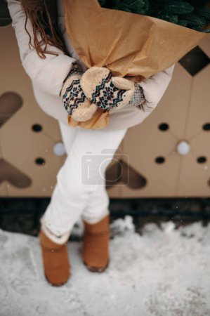 Téléchargez les photos : Fille en tenue d'hiver et gants tient un bouquet d'arbres de Noël dans ses mains. Vue dégagée. Journée enneigée d'hiver - en image libre de droit