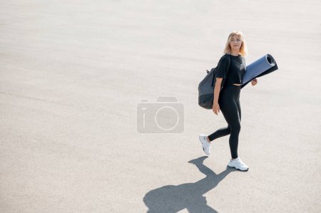 Téléchargez les photos : Jeune femme blonde marchant dans la rue et portant un tapis de yoga tout en se préparant pour la formation en plein air. Concept de mode de vie sain. Espace de copie - en image libre de droit