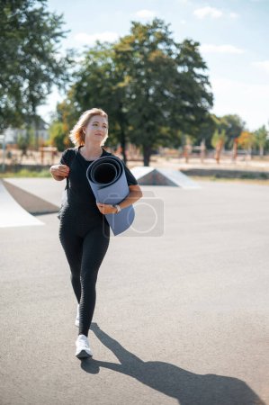 Téléchargez les photos : Joyeuse jeune fille caucasienne tenant tapis de yoga se préparer pour l'entraînement debout à l'extérieur. Femme blonde souriante et marchant dans la rue. Sport et concept sain - en image libre de droit
