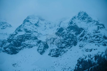 Un paisaje natural de una montaña nevada con rocas y árboles en un día nublado, mostrando un fenómeno geológico con una capa de hielo helada y nubes cúmulos en el cielo azul eléctrico. Morskie Oko, Polonia