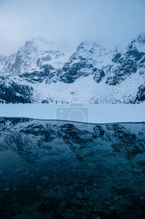 Photo for A natural landscape of a Morskie Oko lake surrounded by snowcovered mountains with a reflection of the mountains in the water under the dusky sky. Tatry, Poland - Royalty Free Image