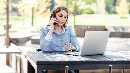 Photo for European businesswoman working outdoors. Beautiful young girl sit at table, talk on mobile phone and use laptop computer. Modern woman lifestyle. Concept of remote and freelance work - Royalty Free Image