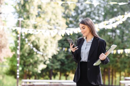 Photo for European businesswoman use mobile phone outdoors. Beautiful focused young girl wear formal suit. Sunny park at daytime. Modern woman lifestyle. Concept of remote and freelance work - Royalty Free Image