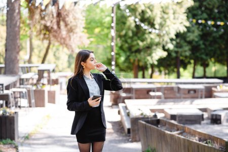 Photo for European businesswoman wait somewhere outdoors. Beautiful positive young girl wear formal suit. Sunny park at daytime. Modern woman lifestyle. Concept of remote and freelance work - Royalty Free Image