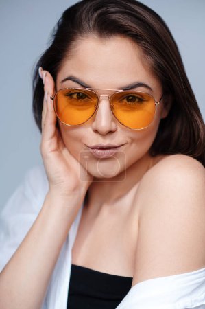Photo for Stylish female model with light natural makeup wearing trendy orange sunglasses against grey background. Glamorous woman posing in studio - Royalty Free Image
