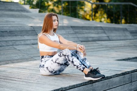 Photo for Young sporty girl in stylish sportswear white top and leggings sitting and rest after trining outdoor on the wooden steps in the park. - Royalty Free Image