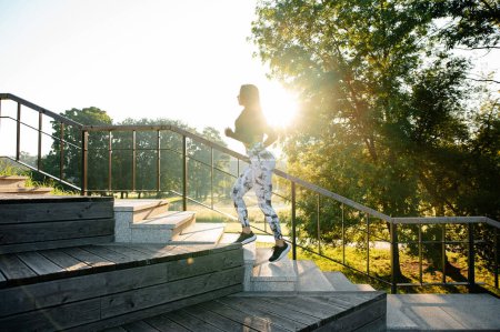 Photo for Young fitness woman in stylish sportswear rashguard and leggings running on stairs up in beautiful green park at sunset. Concept of active and healthy lifestyle - Royalty Free Image