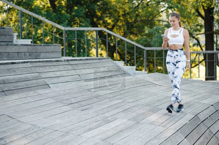Téléchargez les photos : Belle jeune femme sportive marchant avec un smartphone sur la jetée en bois ou des escaliers dans le parc. Smilling fille de remise en forme souriant et regardant au téléphone - en image libre de droit