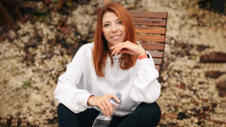 Téléchargez les photos : Jeune femme rousse sportive en tenue de sport assise sur un banc et tenant une bouteille d'eau. Heureuse fille élégante sentant et regardant vers la caméra - en image libre de droit