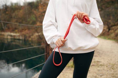 Photo for Sporty girl in a white sweatshirt and black leggings holds a red skipping rope near lake on the park. Cropped view fittnes woman - Royalty Free Image