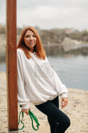 Photo for Portrait of happy red-haired girl in a white sweatshirt and black leggings on the background of the river. Sporty smilling woman holding resistance band and lloking to the camera - Royalty Free Image