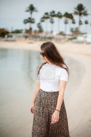 Foto de Mujer caucásica feliz en gafas de sol y ropa elegante caminando en la playa de arena cerca del mar azul. Palmeras y hotel moderno en el fondo. - Imagen libre de derechos