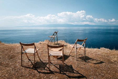 Foto de Cuatro taburetes vacíos están en el borde de las rocas contra el fondo del cielo azul y el mar en Grecia - Imagen libre de derechos