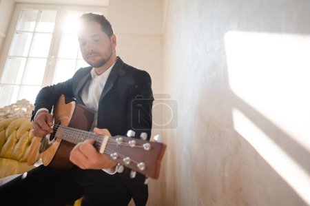 Photo for Low angle view of handsome young beared man playing guitar in the modern hotel room with sunlight. Wedding day - Royalty Free Image