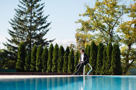 Foto de Guapo novio en traje elegante y con gafas de sol sentado en el borde de una piscina y mirando hacia otro lado en el soleado día de verano. Día de la boda - Imagen libre de derechos