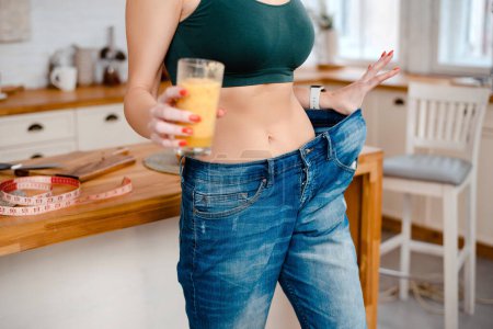 Photo for Close up view of sporty woman posing on camera in oversized jeans and holding glass with vitamin cocktail. Weight loss and diet concept. - Royalty Free Image