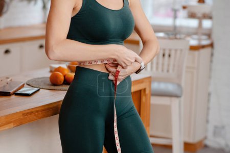 Photo for Cropped view of the sporty woman measuring waist with tape at bright kitchen. Weight loss and diet concept - Royalty Free Image