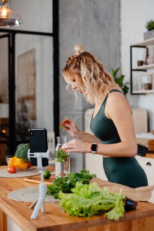 Téléchargez les photos : Blogueur de fitness blonde concentré préparant de la limonade tout en enregistrant une vidéo sur smartphone sur un régime alimentaire sain. Jeune femme en tenue de sport debout sur la cuisine - en image libre de droit
