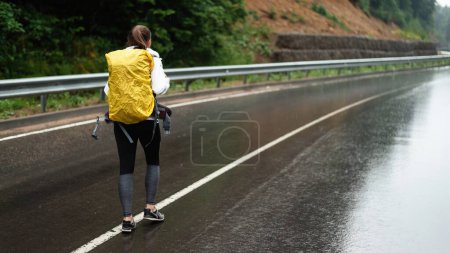 Téléchargez les photos : Vue arrière du randonneur féminin avec sac à dos jaune marchant sur la route asphaltée avec de belles montagnes autour. Concept de personnes et d'activités de loisirs. - en image libre de droit