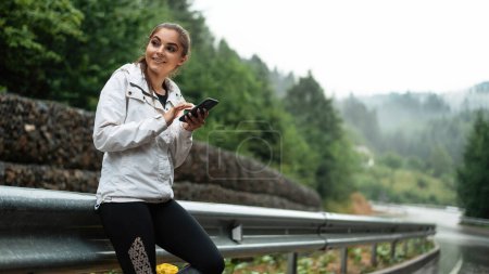Téléchargez les photos : Jeune femme caucasienne vérifiant la direction sur smartphone moderne tout en faisant de la randonnée dans les montagnes seules. Voyageuse debout sur route asphaltée humide avec téléphone portable dans les mains. - en image libre de droit
