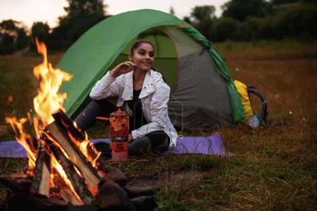 Téléchargez les photos : Happy backpacker woman sitting in entrance of tourist tent at campfire and looking away near forest under sky. Camping at the forest concept - en image libre de droit