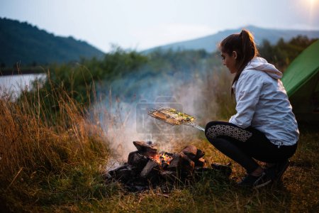 Photo for Young calm woman tourist frying vegetables at the beautiful nature landscape while sitting near the tent by the fire. Travel, tourism, camping concept - Royalty Free Image