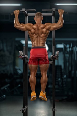 Foto de Un hombre musculoso fuerte con un torso desnudo realiza un estiramiento vertical en la barra, demostrando los músculos de la espalda de forma aislada. Culturista con buenas proporciones, concepto de fuerza y salud - Imagen libre de derechos