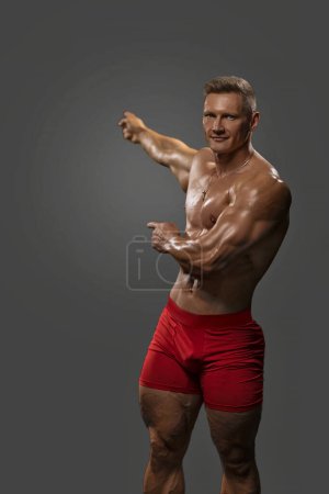 Foto de Un hombre musculoso fuerte sin camisa en pantalones cortos ajustados rojos apunta con ambas manos a un fondo vacío. Fuerza y motivación, llamada a la acción - Imagen libre de derechos