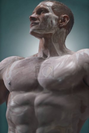 Foto de A strong muscular man with perfect abs is posing in the studio against an empty background. Athletic male body is covered with white clay in the style of an antique statue - Imagen libre de derechos