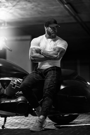 Foto de Sexy guapo atractivo deportivo modelo de fitness musculoso motero empanado en gorra negra y camiseta apretada blanca se sienta en la hermosa motocicleta brutal en el aparcamiento, foto en blanco y negro - Imagen libre de derechos