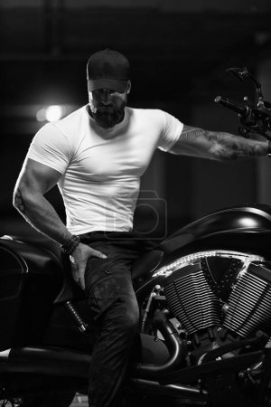 Foto de Sexy guapo atractivo deportivo modelo de fitness musculoso motero empanado en gorra negra y camiseta apretada blanca se sienta en la hermosa motocicleta brutal en el aparcamiento, foto en blanco y negro - Imagen libre de derechos