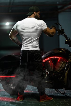 Foto de Sexy guapo atractivo deportivo musculoso modelo de fitness motero empanado con brutal hermosa motocicleta en el estacionamiento posando con la moto con la espalda al marco - Imagen libre de derechos