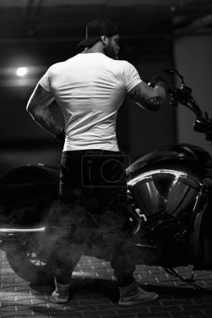 Foto de Sexy guapo atractivo deportivo modelo de fitness musculoso motero empanado con brutal hermosa motocicleta en el estacionamiento posando con una moto con la espalda al marco, foto en blanco y negro - Imagen libre de derechos