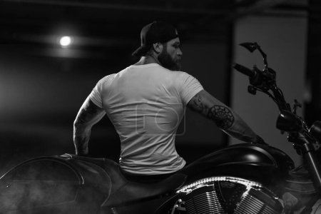 Foto de Sexy guapo atractivo deportivo modelo de fitness musculoso motero empanado con brutal hermosa motocicleta en el estacionamiento posando con una moto con la espalda al marco, foto en blanco y negro - Imagen libre de derechos