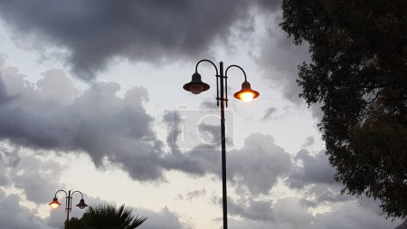 Eine Straßenlaterne hängt an einem Mast vor dem Hintergrund eines Baumes mit Wolken und blauem Himmel. Das atmosphärische Gas erhellt die Kumuluswolken bei windigem Wetter