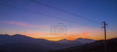 Téléchargez les photos : Une image floue capturant un ciel rouge le matin avec la silhouette d'une chaîne de montagnes et des lignes électriques au premier plan, créant un paysage naturel tranquille - en image libre de droit