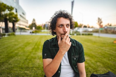 Téléchargez les photos : Un homme aux cheveux bouclés s'assoit sur un parc, fume un cigare et se repose - en image libre de droit