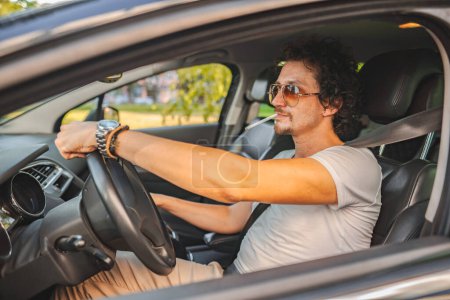 Photo for Handsome man with curly hair and sunglasses, drives a car and smokes a cigarette. - Royalty Free Image