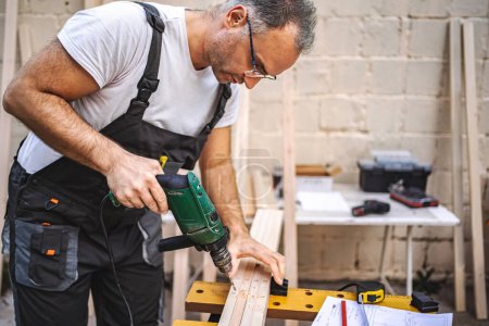 Téléchargez les photos : Mature mâle charpentier à l'aide de tournevis électrique sur une planche en bois à l'extérieur. - en image libre de droit