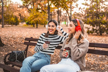 Téléchargez les photos : Deux femmes sont assises sur un banc en bois dans un parc, écoutent de la musique à l'aide d'écouteurs et aiment boire du café et socialiser.. - en image libre de droit