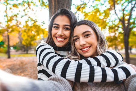 Téléchargez les photos : Deux femmes d'ethnies différentes s'embrassent chaleureusement dans un parc verdoyant. Ils sont debout sur un chemin entouré d'arbres tout en partageant un câlin significatif. - en image libre de droit