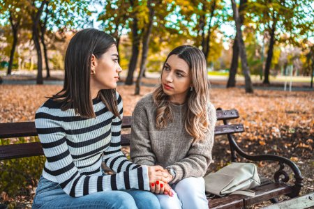 Two women are sitting on a park bench. They are facing each other, looking at each other, holding hands, comforting each other and giving each other comfort and support.