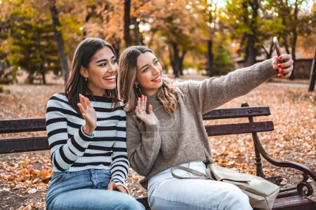 Foto de Dos mujeres están sentadas en un banco de madera en un parque. Se están tomando una selfie y disfrutando de la compañía de los demás. Los árboles y la hierba los rodean, creando un ambiente tranquilo. - Imagen libre de derechos